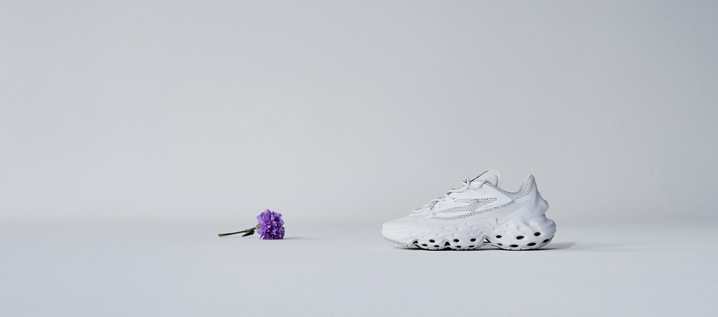A picture of a purple flower on the left and a Seed.One shoe in the White Lily colorway from the lateral view on the right facing each other in a total position