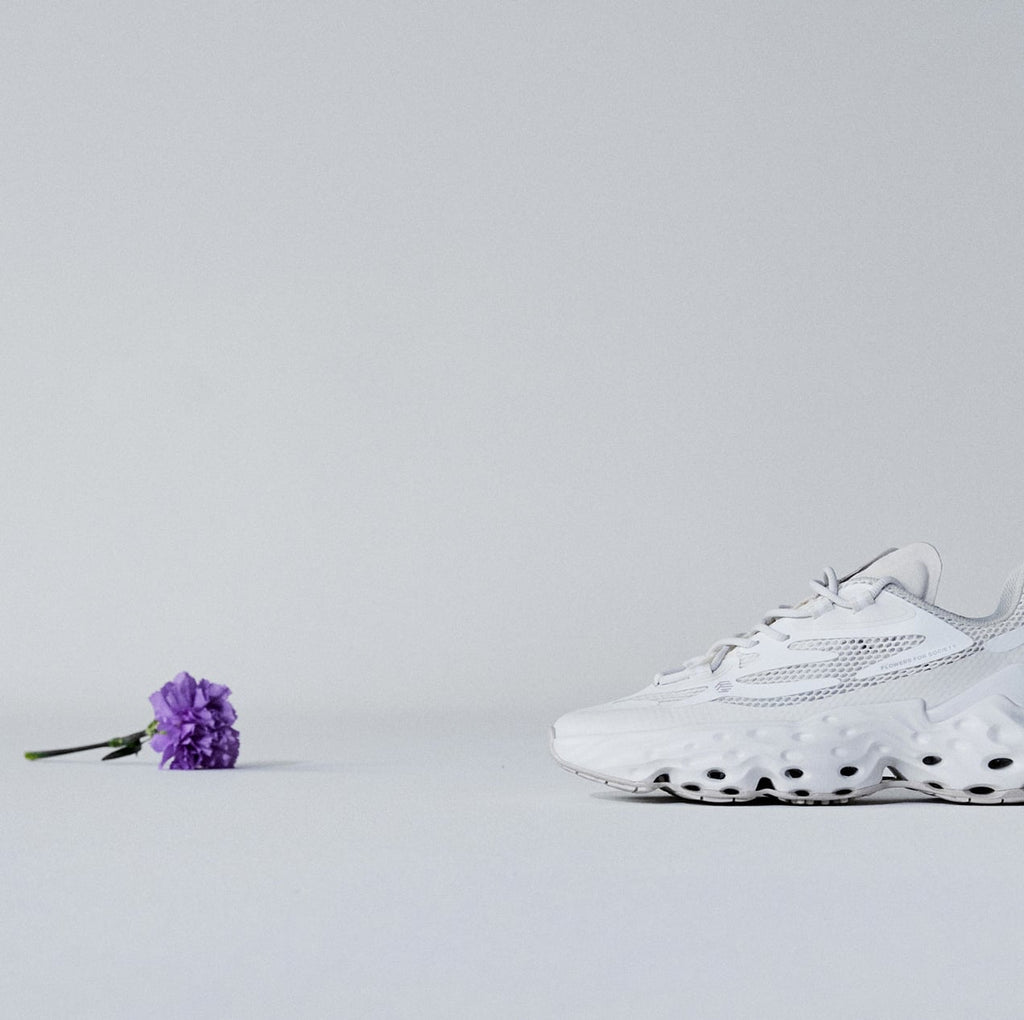A half total picture of a purple flower on the left and a Seed.One shoe in the White Lily colorway from the lateral view on the right facing each other