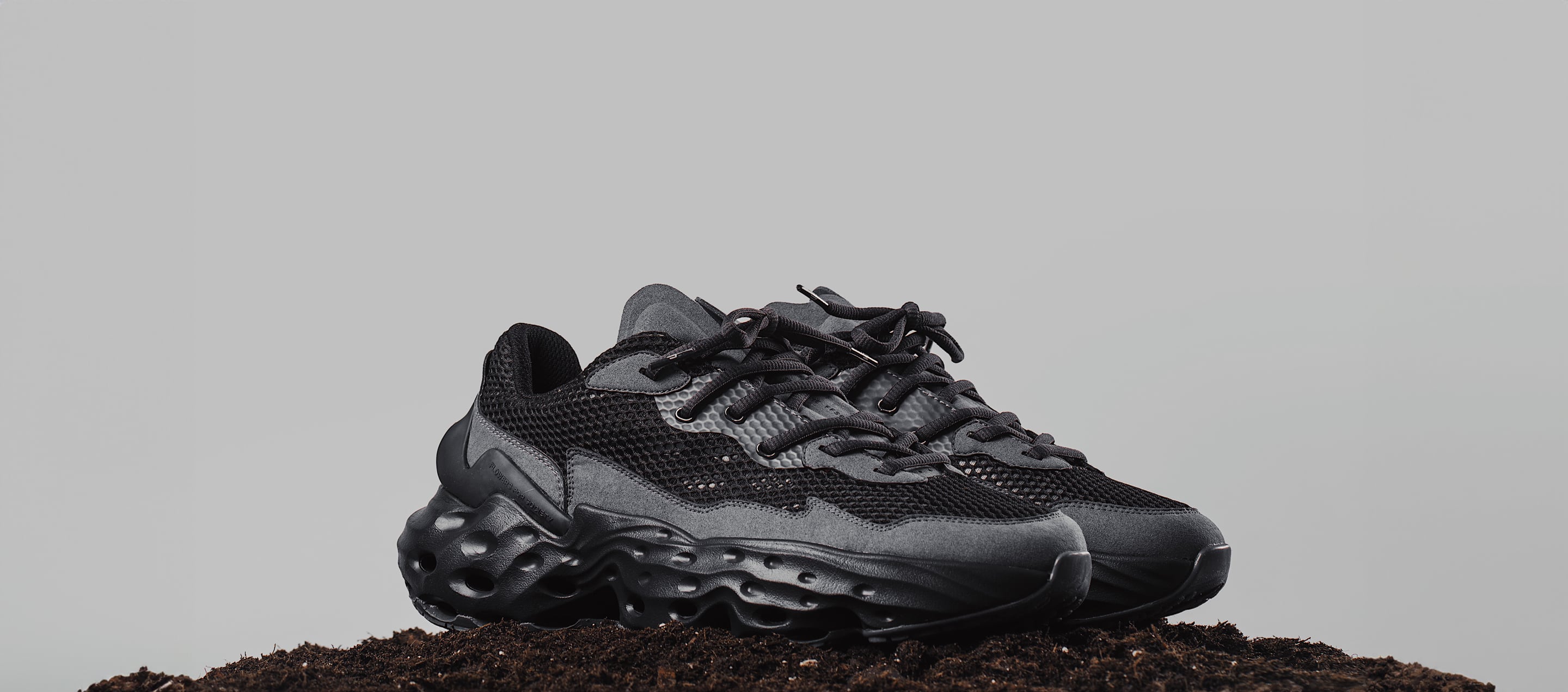 A campaign picture of the shoe Seed.One in the mainly black and dark gray colorway called Black Mud from the diagonal view standing on a pile of soil on a gray background