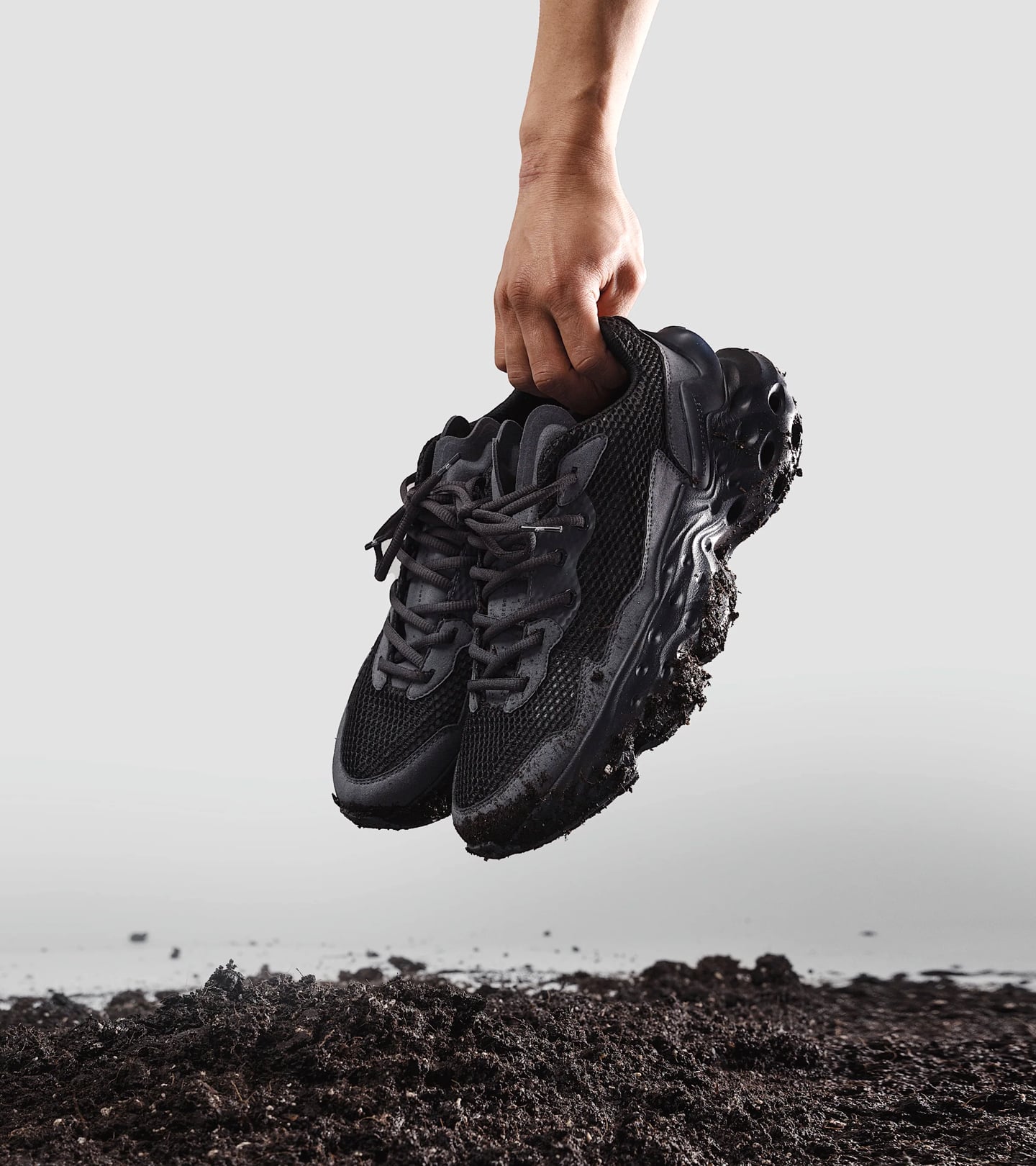 A hand hanging down from above holding a pair of Seed.One Black Mud above a pile of soil on a gray background