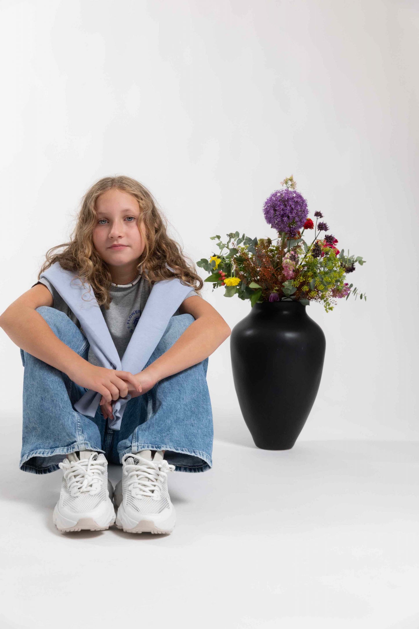 Flowers for Society basic kids hoodie blue front view worn by model over the shoulders sitting on the ground next to a flower vase