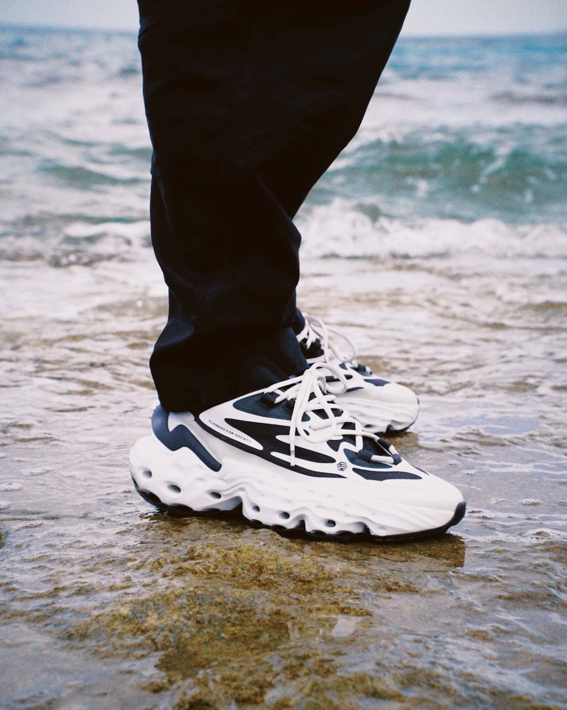 Flowers For Society Sneaker Seed.One Orca black white lateral worn by model standing in shallow water